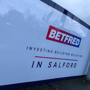 A large construction or advertising panel featuring the Betfred logo in red, white, and blue, with the words "INVESTING • BUILDING • BELIEVING" and "IN SALFORD" written underneath, situated beside a metal pole and a cloudy sky reflected on the panel.