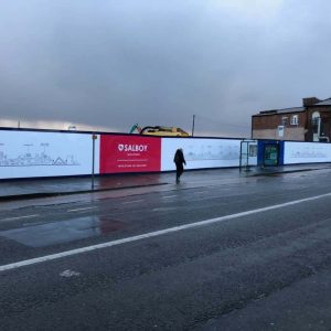 A long construction barricade with illuminated advertisements, including a prominent red "Salboy" banner, against a cloudy, overcast sky. There are a few pedestrians walking along the wet sidewalk, and old brick buildings are visible in the background.