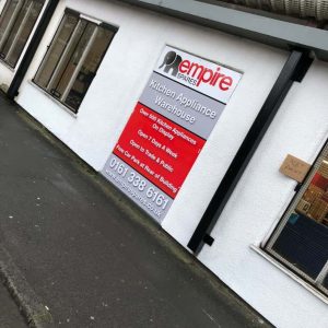 A large shop fascia sign for "Empire Spares," mounted on the white exterior wall of a building. The sign advertises kitchen appliances and warehouse stock, with contact details and website information.