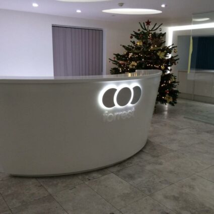 A modern reception desk with illuminated signage on the front, featuring the logo "COO" and the word "forrest" beneath it, in a clean, contemporary office space with a decorated Christmas tree in the background.