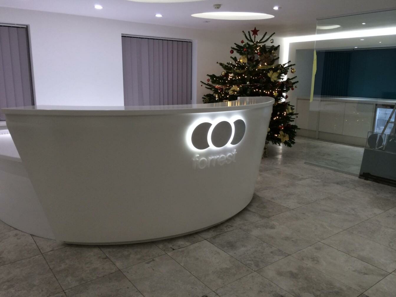A modern reception desk with illuminated signage on the front, featuring the logo "COO" and the word "forrest" beneath it, in a clean, contemporary office space with a decorated Christmas tree in the background.