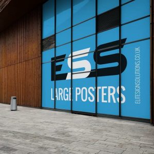 A large outdoor sign with a bright blue background displaying the text "LARGE POSTERS" and a website URL, mounted on the exterior wall of a modern building with wooden and glass architectural elements.