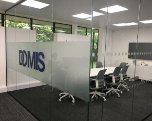 Glass wall partition with the "DOMIS" logo in blue and grey, inside a modern office room.