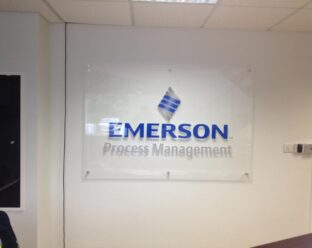 A clear acrylic wall sign with the Emerson Process Management logo in blue and grey, mounted on a white wall.