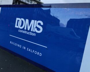 Construction hoarding with the logo "DOMIS construction" and the tagline "BUILDING IN SALFORD" on a blue background.
