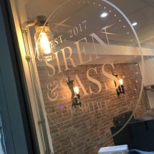 A glass window with the logo and text "Siren & Sass" printed in white. The logo includes "EST. 2017" above the name and "CHESTER" below, all enclosed within a circular border. Inside, a warm Edison-style light bulb is mounted on a metal fixture, illuminating the space behind the window, which features a brick wall with two wall-mounted candle-style sconces. There are also some items on a table, including a box, a cup, and a piece of clothing.