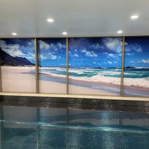 Colorful mural of a beach scene with blue skies, white clouds, and ocean waves, displayed on a large window inside a swimming pool area.
