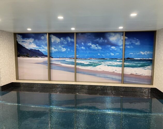 Colorful mural of a beach scene with blue skies, white clouds, and ocean waves, displayed on a large window inside a swimming pool area.