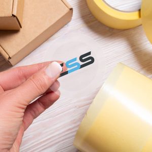 A hand peeling a transparent vinyl sticker with a colorful logo from a sheet, surrounded by rolls of yellow tape and a cardboard box on a light wooden surface.