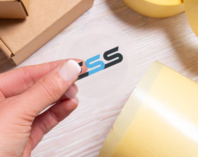 A hand peeling a transparent vinyl sticker with a colorful logo from a sheet, surrounded by rolls of yellow tape and a cardboard box on a light wooden surface.