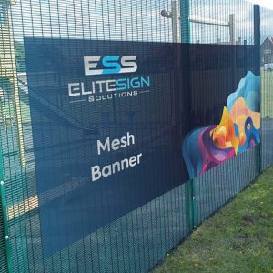 A large mesh banner with colourful abstract artwork on the right side, mounted outdoors on a metal fence.